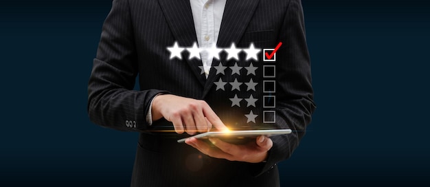 Photo businessman hand touching on digital mobile tablet with five star rating to review good feedback on checklist box on dark background, online marketing, good experience, customer feedback concept