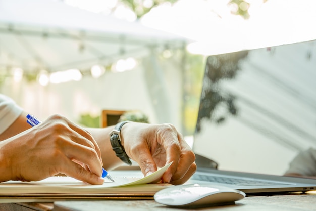 Mano di uomo d'affari che scrive su carta del notebook con un computer portatile sul tavolo.