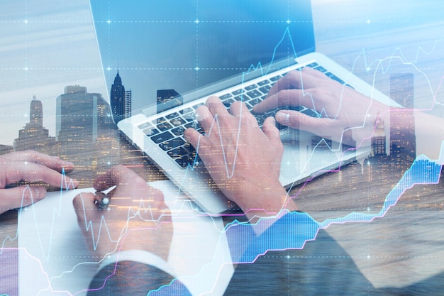 Businessman hands writing in planner, businesswoman hands typing on laptop over cityscape background with double exposure of blue graph.