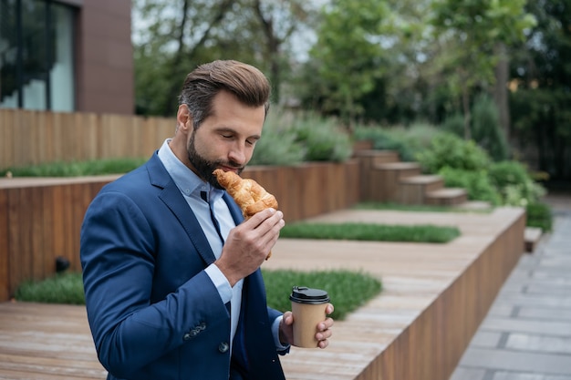 路上でクロワッサンを食べるコーヒーのカップを保持しているビジネスマンコーヒー休憩ランチ時間の概念