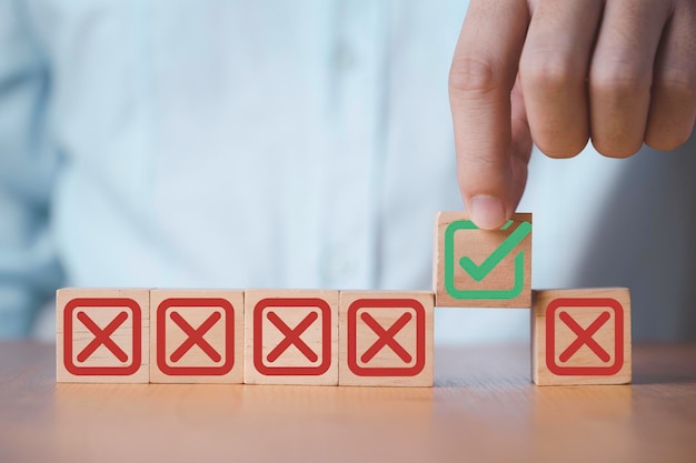 Businessman holding green correct sign mark among Red Cross mark which print screen on wooden cube block for approve and reject business proposal concept