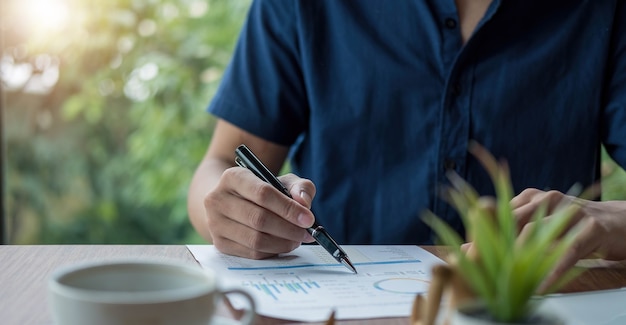 Uomo d'affari che tiene la penna e indica il riepilogo del grafico cartaceo analizzando il rapporto annuale di attività con l'utilizzo del computer portatile alla scrivania dell'ufficio della stanza.