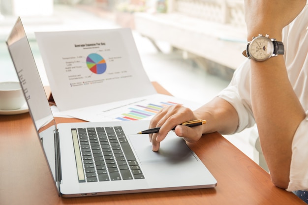 Businessman holding pen working on laptop with financial graph report on table.