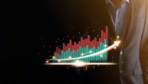 Businessman holding and showing a growing virtual hologram of statistics, graph and chart with arrow up on dark background. Stock market. Business growth, planing and strategy concept.