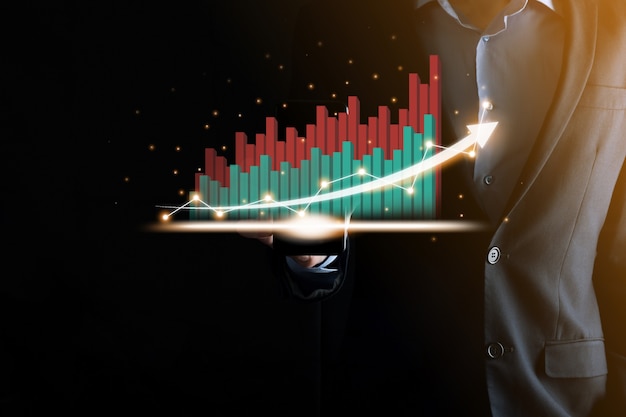 Businessman holding and showing a growing virtual hologram of statistics, graph and chart with arrow up on dark background. Stock market. Business growth, planing and strategy concept.