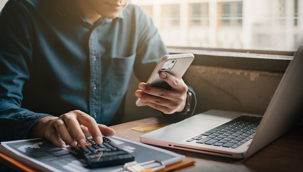 Businessman holding smartphone and computer are calculating on calculator investment costs in hand at home office.