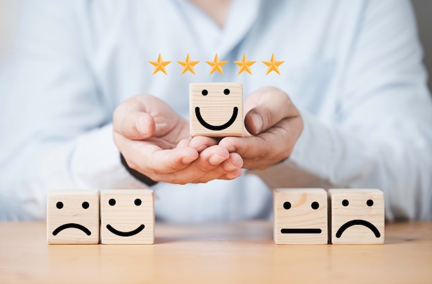 Businessman holding smile face on wooden cube block with other\
face emotion for excellent evaluation and customer satisfaction\
concept