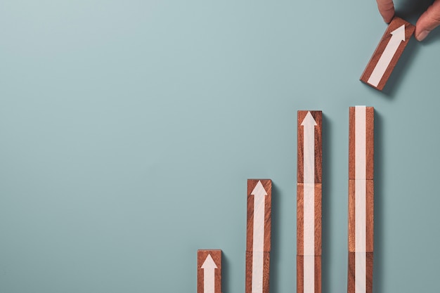 Businessman holding and stacking white up arrow which print screen on wooden block on blue background , Business profit growth concept.