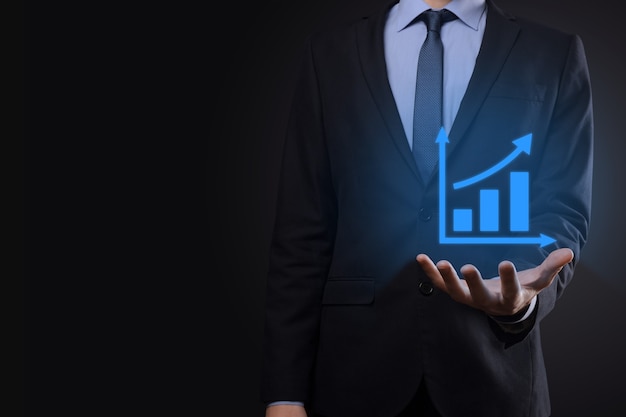 Businessman holding tablet and showing a growing virtual hologram of statistics, graph and chart with arrow up on dark background. Stock market. Business growth, planing and strategy concept.