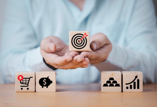 Businessman holding target board which printing on wooden cube\
block with icons include shopping trolley piggy saving gold and\
graph for set up business objective investment goal concept