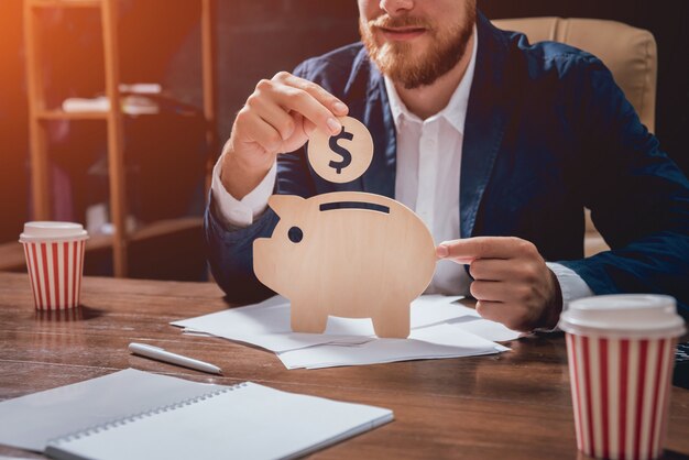 Businessman holds a coin under wooden piggy.