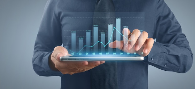 Businessman holds a tablet with growing graph hologram on grey background