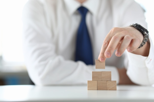 Businessman holds wooden cube in his hand and builds a pyramid.