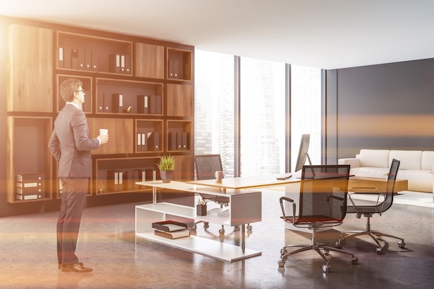 Businessman in interior of ceo office with gray walls, concrete floor, white and wooden computer table and dark wooden shelves with folders. Beige sofa near coffee table. Toned image