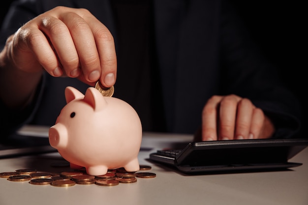 Businessman is counting profit on a calculator in office, piggy bank with coins at desk. Economy and management financial concept