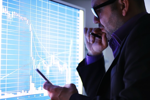 A businessman is looking at a graph on a monitor. An exchange broker evaluates stock market trends. A man with glasses in front of a curve of the dynamics of the economy.