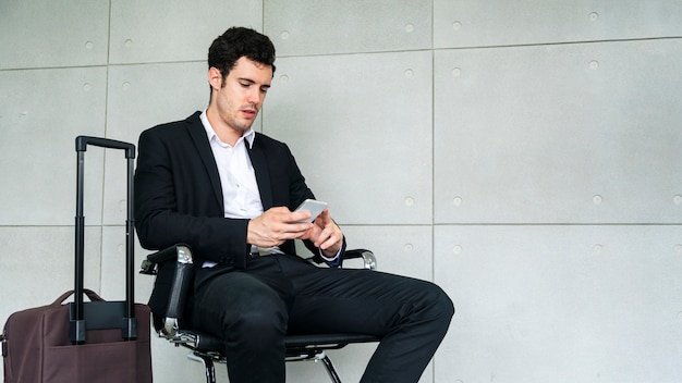 Businessman is sitting on a chair waiting with a suitcase and smartphone