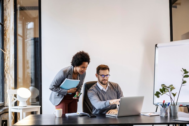 A businessman is sitting at the office and pointing at the laptop while talking to a manager