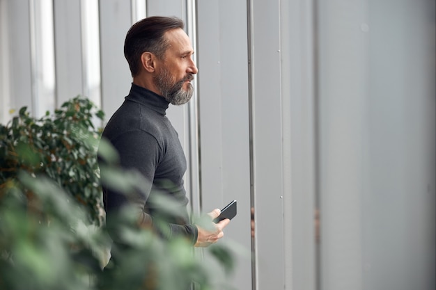 Businessman is standing near big windows in office space and holding smartphone