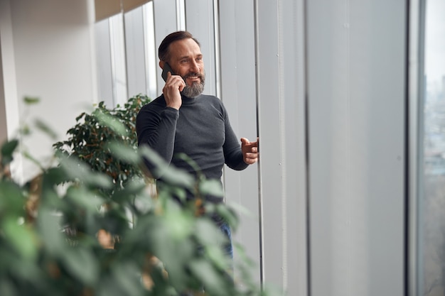 Businessman is standing near big windows in office space and holding smartphone