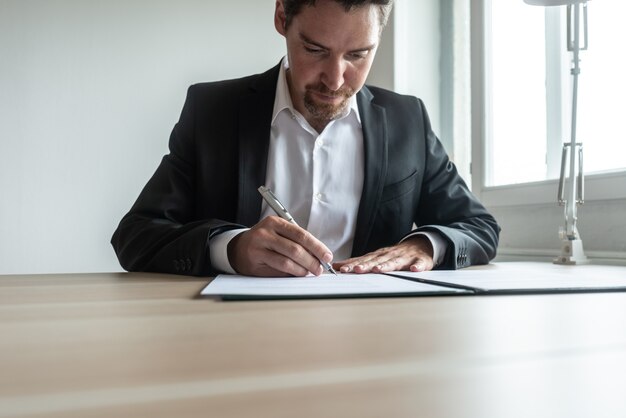 Бизнесмен или юрист, сидя за своим офисным столом, подписывая документ или контракт в папке.