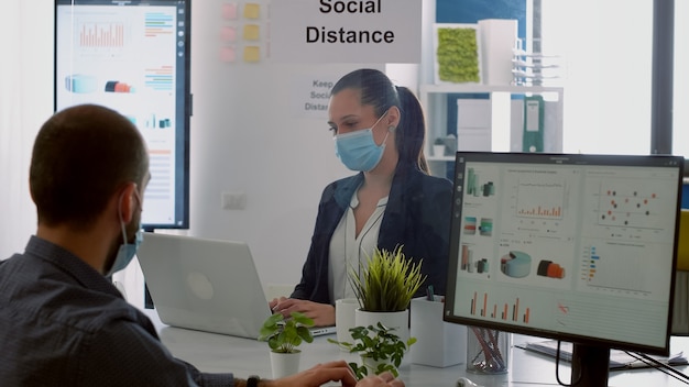 Businessman looking at financial graphs on computer display while talking with coworker sitting in company office. Colleagues with face masks keeping social distancing to prevent covid19 disease