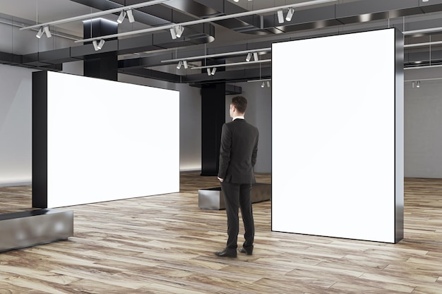 businessman looks at blank white partitions with place for advertising poster or marketing campaign in stylish gallery hall with metallic benches on wooden floor and grey wall background mockup