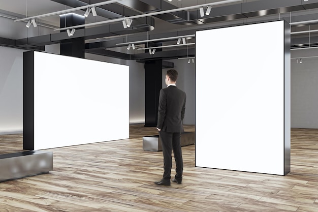 Photo businessman looks at blank white partitions with place for advertising poster or marketing campaign in stylish gallery hall with metallic benches on wooden floor and grey wall background mockup