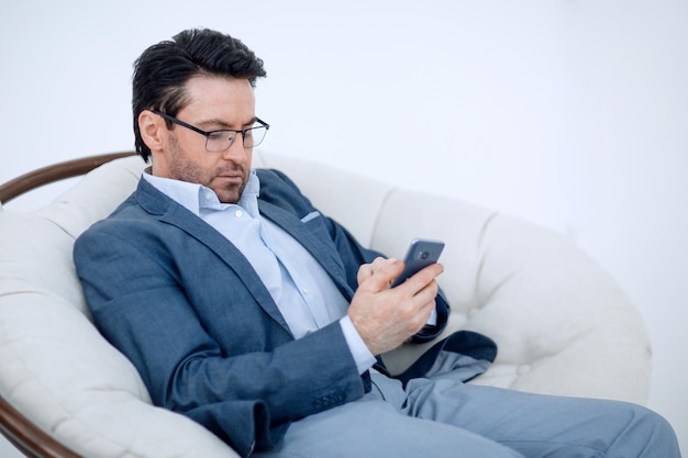 Бизнесмен смотрит на экран смартфона, сидя в удобном кресле