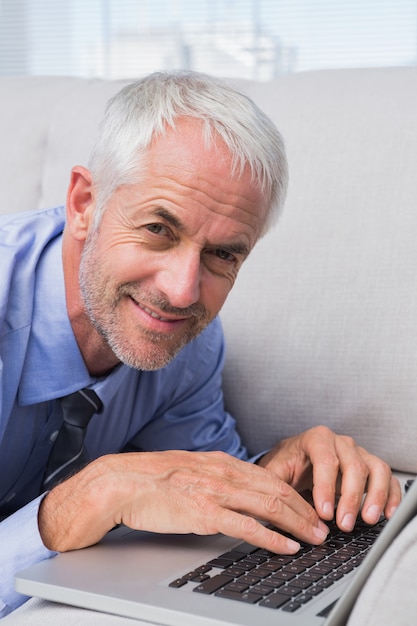 Businessman lying on couch with laptop and smiling at camera