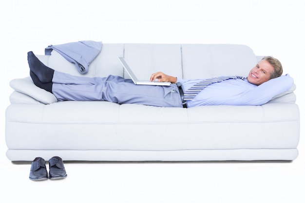  Businessman lying on sofa using his laptop smiling at camera