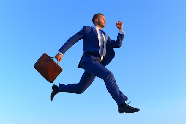 Photo businessman makes big step up on career ladder business and success concept project manager with strict face expression man in blue formal suit holds brown case and jumps up on blue sky background