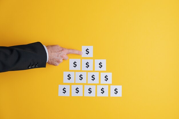 Businessman making a pyramid shape foundation of white post it papers with dollar sign on them in a conceptual image of profit and power. over yellow background with copy space