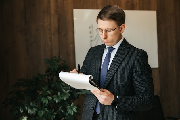 眼鏡をかけたビジネスマンの男性は、オフィスで書類を書き留めます。