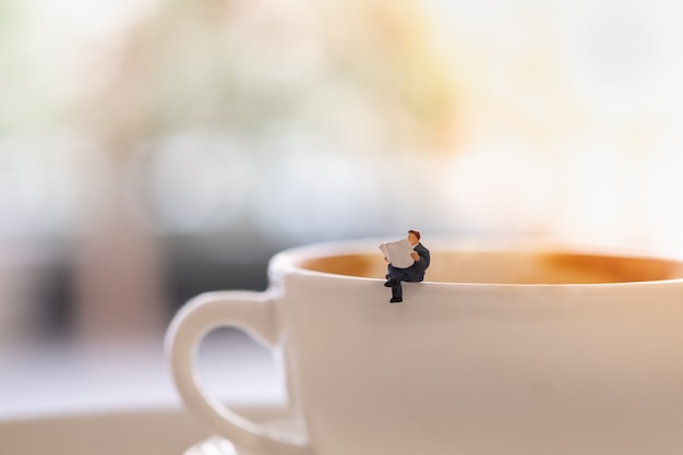 businessman miniature mini people figures sitting and read a newspaper on the cup of hot coffee.