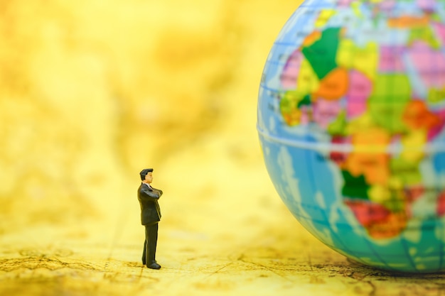 Businessman miniature people figure standing on map and looking to mini world ball on map.