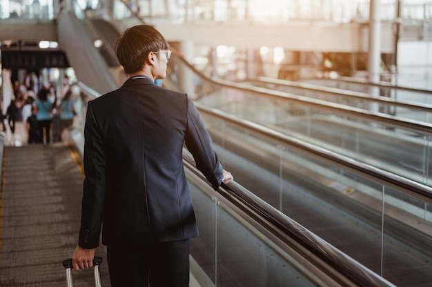 空港で荷物を持ってチェックインするためにエスカレーターでターミナルゲートに移動するビジネスマン。