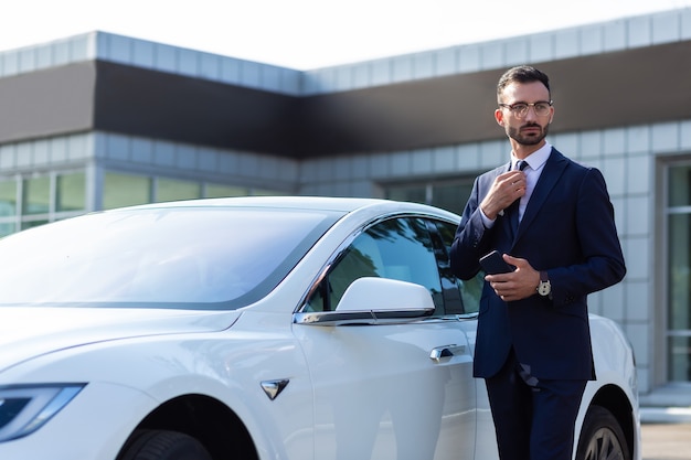 Uomo d'affari vicino all'auto. elegante giovane uomo d'affari in piedi vicino a un'auto bianca al mattino