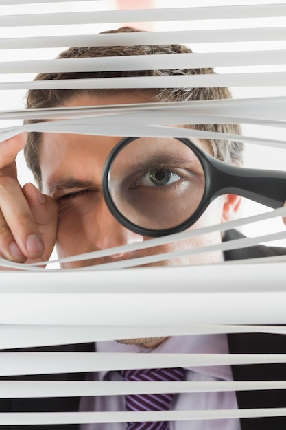 Businessman peeking through blinds with magnifying glass