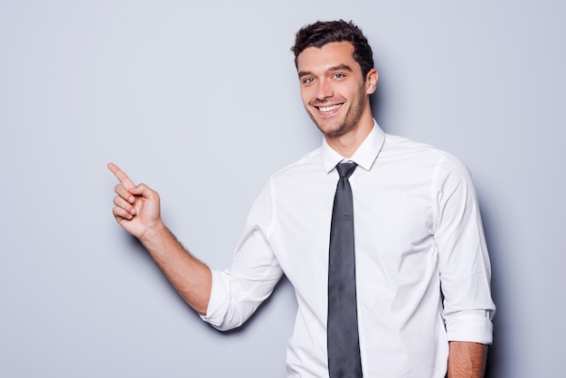 Бизнесмен указывая копией пространства. Счастливый молодой человек в рубашке и галстуке смотрит в камеру и улыбается, стоя на сером фоне и указывая копией пространства