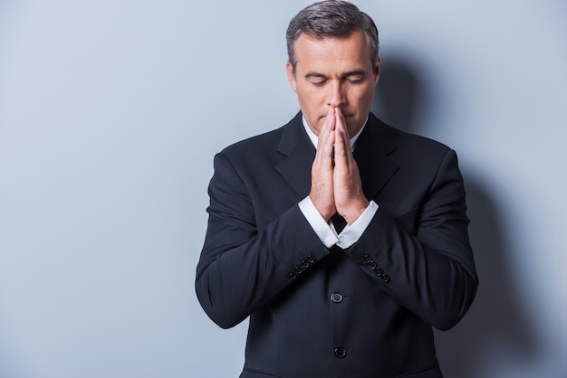 Businessman praying. Thoughtful mature man in formalwear holding hands clasped near face and keeping eyes closed while standing against grey background