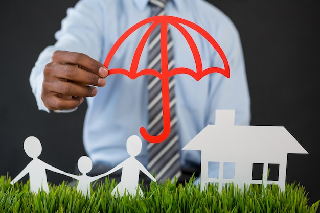 Businessman protecting paper cut out family, house and car with umbrella