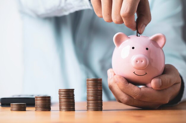 Businessman putting coin to pink piggy bank with increasing coins stacking on table Money saving and deposit for investment to get profit and dividend concept