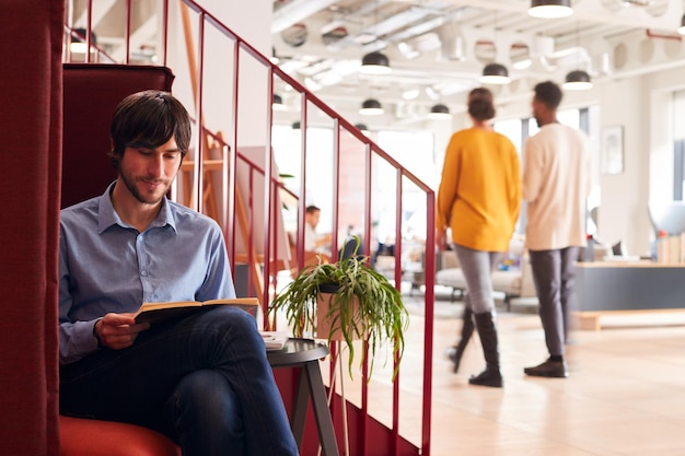 Businessman Reading Book Sitting By Stairs In Modern Open Plan Office