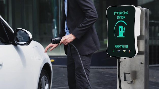 Бизнесмен заряжает свой электромобиль на зарядной станции в центре города или на общественной автостоянке. Экологически чистый перезаряжаемый автомобиль, использующий альтернативную чистую энергию в городском образе жизни.