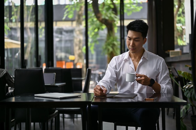 A businessman remote working at a coffee shop sipping coffee and taking notes on his notebook