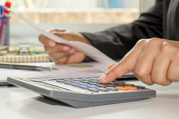 ビジネスマンの手は、電卓を押して、財務作業を行い、ホームオフィスでの経費についてテーブルで計算しています。