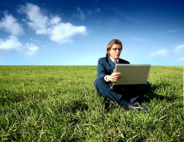 Бизнесмен, сидя на траве поля работает с ноутбуком