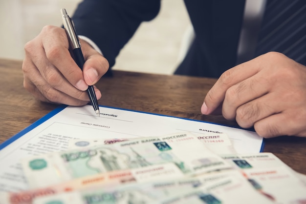 Бизнесмен подписывает договор контрактной бумаги с деньгами, российские рублевые купюры, на столе
