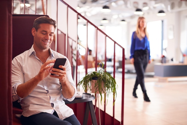 現代のオープンプランのオフィスで階段のそばに座っているビジネスマンが携帯電話でメッセージをチェック